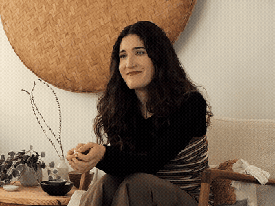 A Woman Sits In A Chair In Front Of A Wicker Wall Hanging GIF