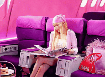 A Woman Sits In A Purple Chair Reading A Magazine While A Red Dutch Bag Sits Next To Her GIF