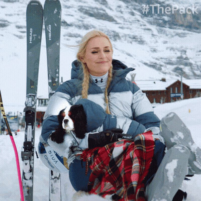 A Woman Sits In The Snow Holding A Dog And A Pair Of Skis That Say Head GIF