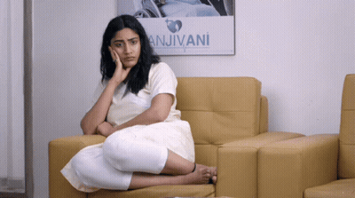 A Woman Sits On A Couch In Front Of A Poster That Says Anjivani GIF