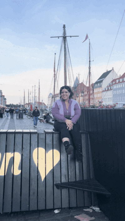 A Woman Sits On A Fence With A Heart Painted On It In Front Of A Boat GIF
