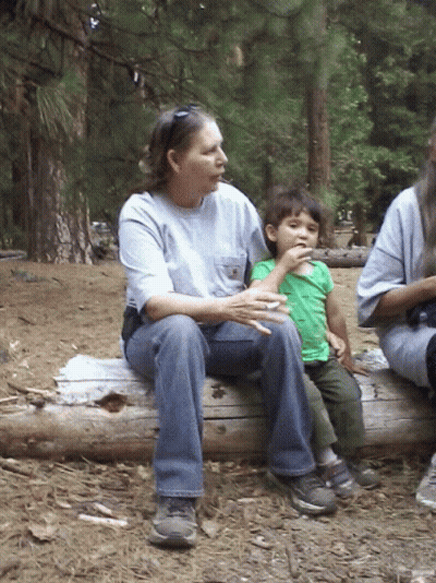 A Woman Sits On A Log Holding A Child Who Is Eating Something GIF