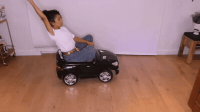 A Woman Sits On A Small Black Toy Car With Her Arms Outstretched GIF