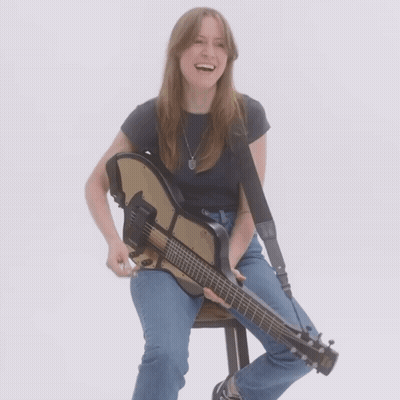 A Woman Sits On A Stool Holding A Guitar And Smiling GIF