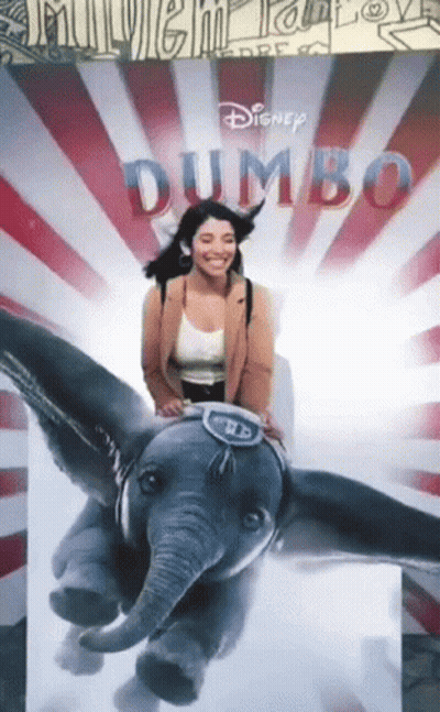 A Woman Sits On A Stuffed Elephant In Front Of A Dumbo Poster GIF