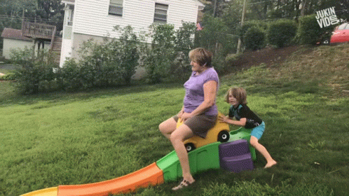A Woman Sits On A Toy Car While A Boy Pushes It Down A Hill GIF