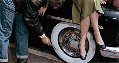 A Woman Sits On The Back Of A Car While A Man Checks The Tire GIF