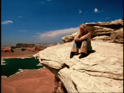 A Woman Sits On The Edge Of A Rocky Cliff Overlooking A Lake GIF