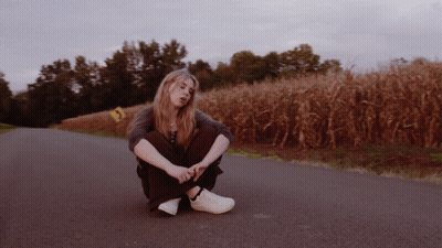 A Woman Sits On The Side Of A Road In Front Of A Cornfield GIF