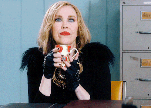 A Woman Sitting At A Desk Holding A Cup With A Label That Says Gold GIF