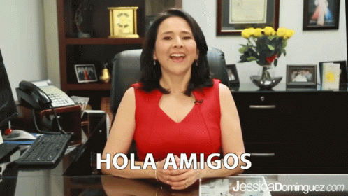 A Woman Sitting At A Desk With Hola Amigos Written On The Top GIF