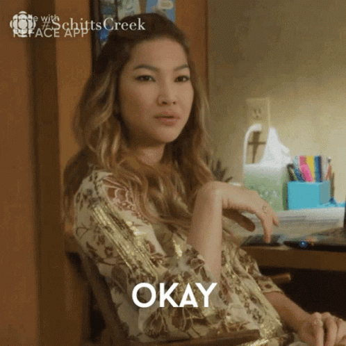 A Woman Sitting At A Desk With Okay Written On Her Shirt GIF