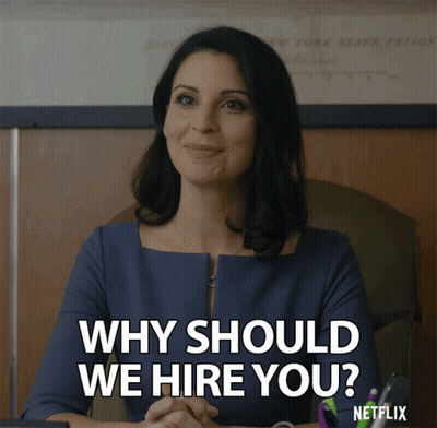 A Woman Sitting At A Desk With The Words " Why Should We Hire You " GIF
