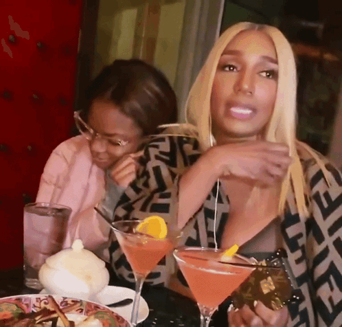 A Woman Sitting At A Table Holding A Martini With A Slice Of Orange In It GIF