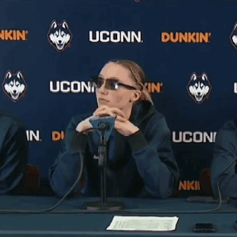 A Woman Sitting At A Table In Front Of A Wall That Says Uconn Dunkin GIF