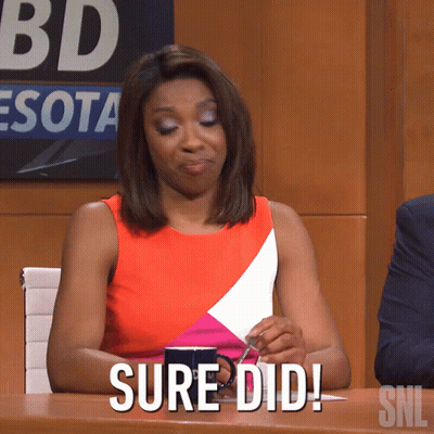 A Woman Sitting At A Table With A Cup Of Coffee And A Sign That Says Snl GIF