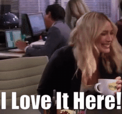 A Woman Sitting At A Table With A Cup Of Coffee And The Words " I Love It Here " Above Her GIF