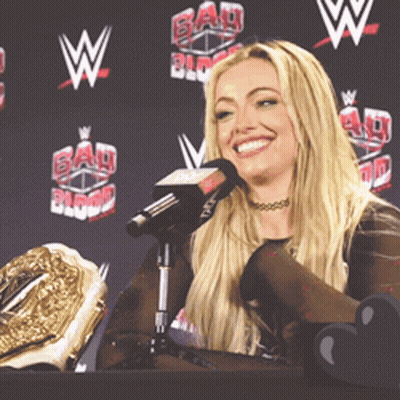 A Woman Sitting At A Table With A Microphone In Front Of A Wall With Bad Blood Written On It GIF