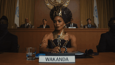 A Woman Sitting At A Table With A Sign That Says Wakanda On It GIF