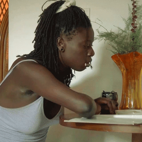 A Woman Sitting At A Table With A Vase Of Flowers In The Background GIF