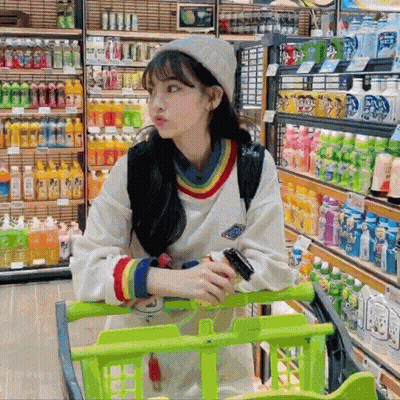 A Woman Sitting In A Shopping Cart In A Store With Bottles Of Soda On Shelves GIF