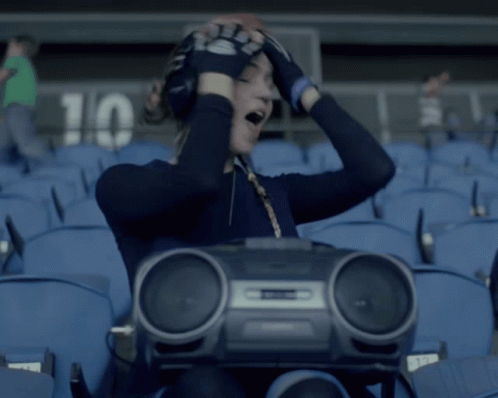 A Woman Sitting In A Stadium Holding A Boombox With The Number 10 On The Wall Behind Her GIF
