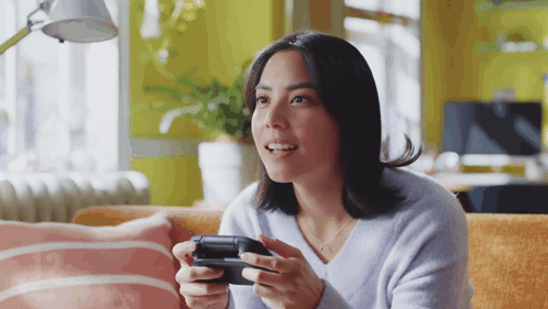 A Woman Sitting On A Couch Holding A Video Game Controller GIF