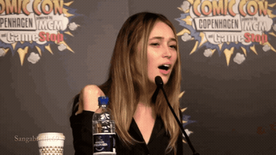A Woman Speaking Into A Microphone In Front Of A Comic Con Banner GIF