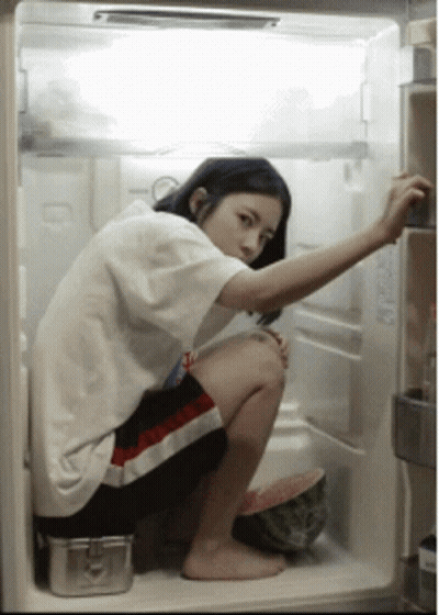 A Woman Squatting In An Open Refrigerator With A Watermelon In The Fridge GIF