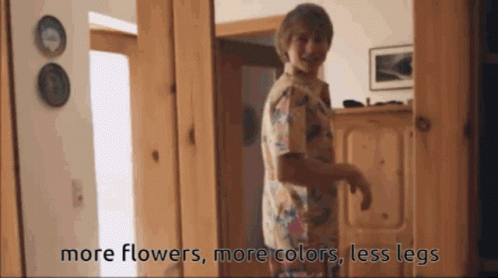 A Woman Standing In A Hallway With The Words More Flowers More Colors Less Legs On The Bottom GIF