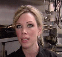 A Woman Standing In A Kitchen With Pots And Pans Hanging On The Wall Behind Her GIF