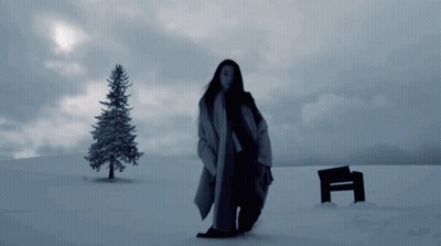 A Woman Standing In A Snowy Field With A Bench And A Tree In The Background GIF