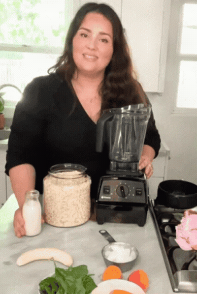 A Woman Standing In Front Of A Blender With A Bottle Of Milk On The Counter GIF