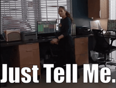 A Woman Standing In Front Of A Desk With The Words Just Tell Me Written Above Her GIF