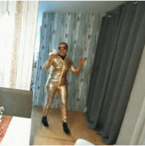 A Woman Standing In Front Of A Mirror Wearing A Gold Jumpsuit GIF