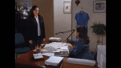 A Woman Standing Next To A Woman Sitting At A Desk In An Office . GIF