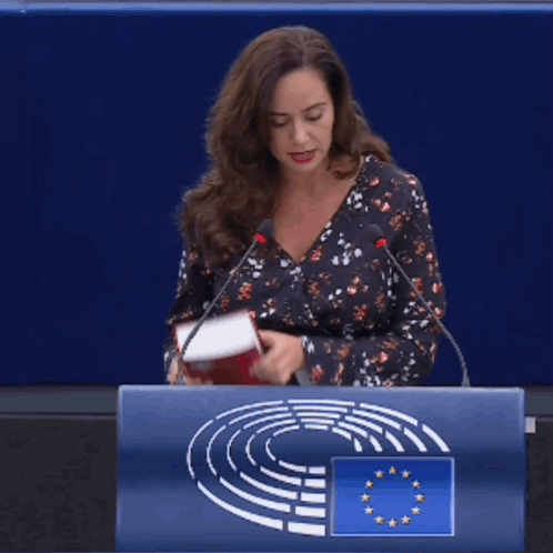 A Woman Stands At A Podium Holding A Red Book That Says Un Code Rural Francais GIF
