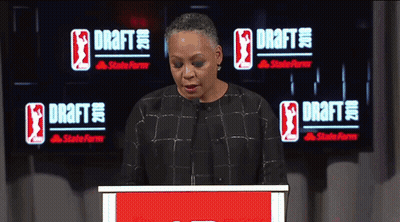 A Woman Stands At A Podium In Front Of A Large Screen That Says Draft GIF