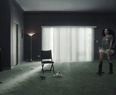 A Woman Stands In A Dark Room With A Chair And A Lamp GIF