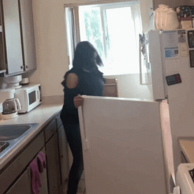 A Woman Stands In A Kitchen Holding A White Refrigerator GIF