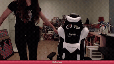 A Woman Stands In Front Of A Gaming Chair That Says T. Jud I Lay-Liv GIF