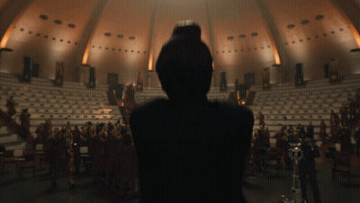 A Woman Stands In Front Of A Large Audience In A Dark Auditorium GIF