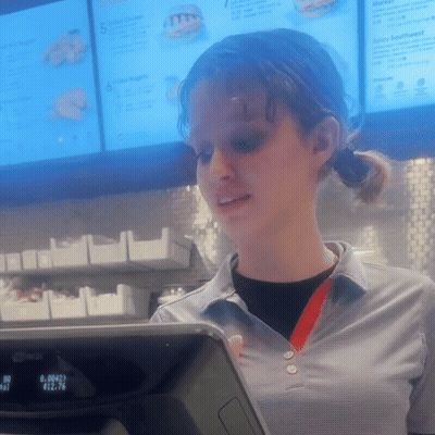 A Woman Stands In Front Of A Mcdonald 'S Checkout Counter With A Menu Behind Her GIF