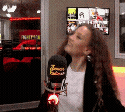 A Woman Stands In Front Of A Microphone That Says Zoomer Radio.com GIF