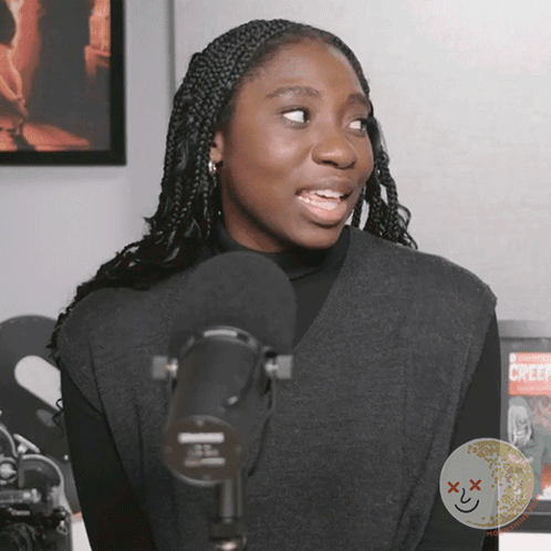 A Woman Stands In Front Of A Microphone With A Creepy Face On The Bottom Right Corner GIF