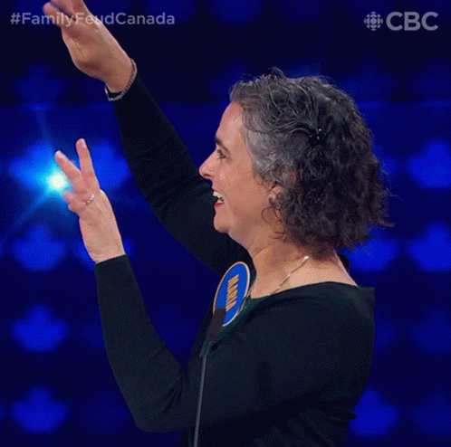 A Woman Stands In Front Of A Microphone With Her Arms In The Air And The Words Family Feud Canada Below Her GIF