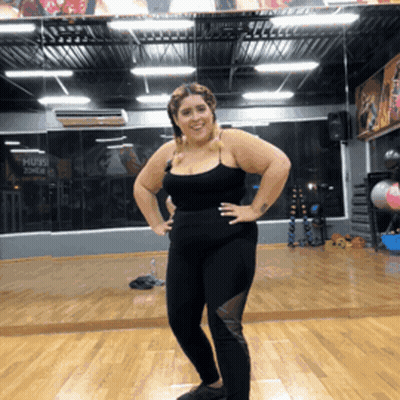 A Woman Stands In Front Of A Mirror In A Gym With A Sign On The Wall That Says ' Hugs ' GIF