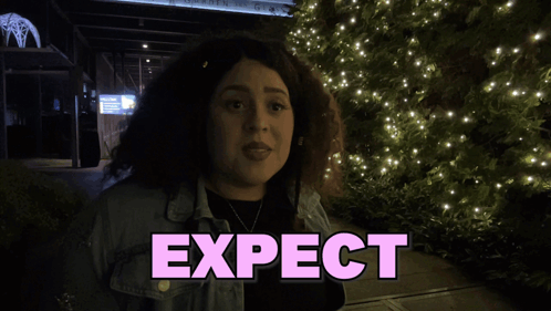A Woman Stands In Front Of A Sign That Says Expect Shagarita GIF