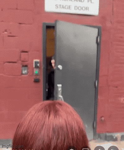 A Woman Stands In Front Of A Stage Door That Is Labeled 212 Ashland Pl GIF