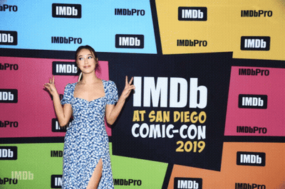 A Woman Stands In Front Of A Wall That Says Imdb At San Diego Comic Con GIF